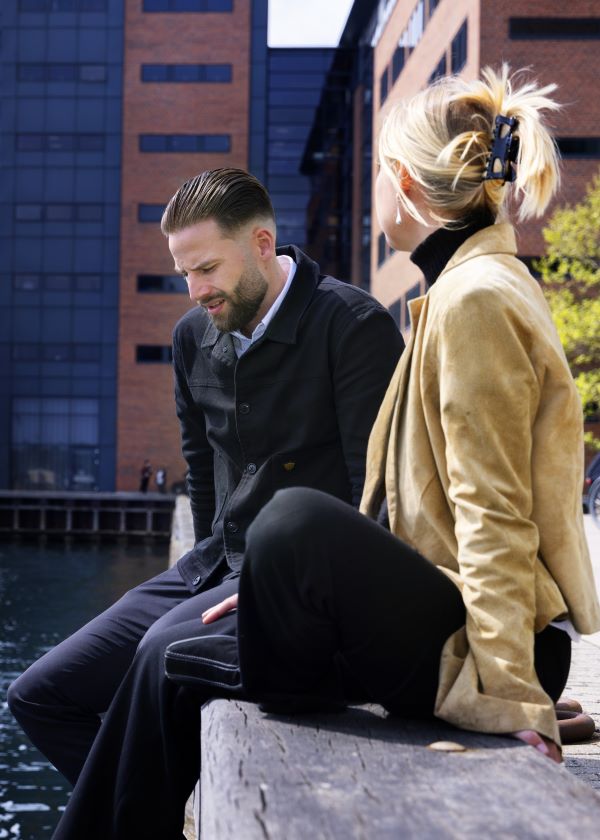 to personer sidder ved en kajkant - personen i baggrunden kigger ned og personen i forgrunden vender mod vedkommende