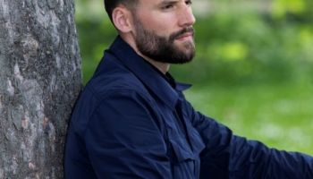 en person i mørkt tøj sidder lænet op imod et træ og kigger ud i billedets højre side