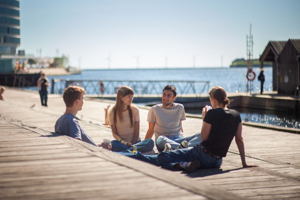fire personer sidder i en cirkel ved vandet i et bymiljø