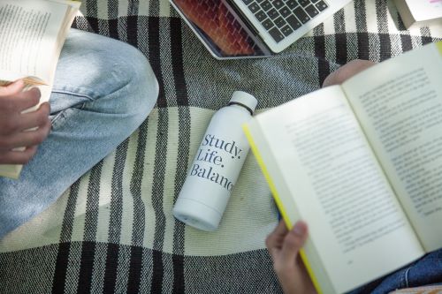 en vandflaske på et tæppe omringet af bøger, ben og computere. på flasken står "study. life. balance."