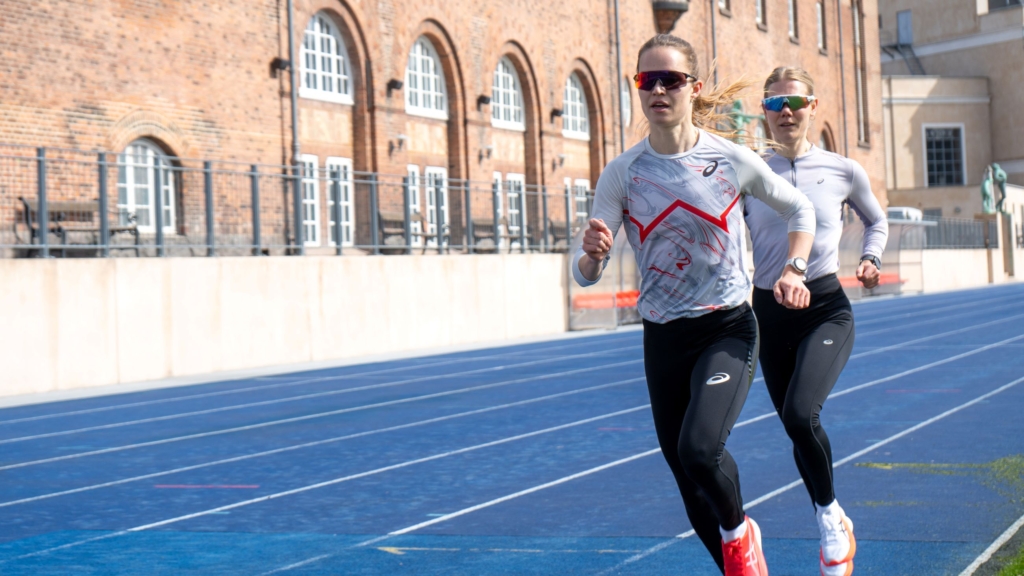 to personer på en blå løbebane. I baggrunden en murstensbygning med vinduer og en hvid mur med jerngitter foran.