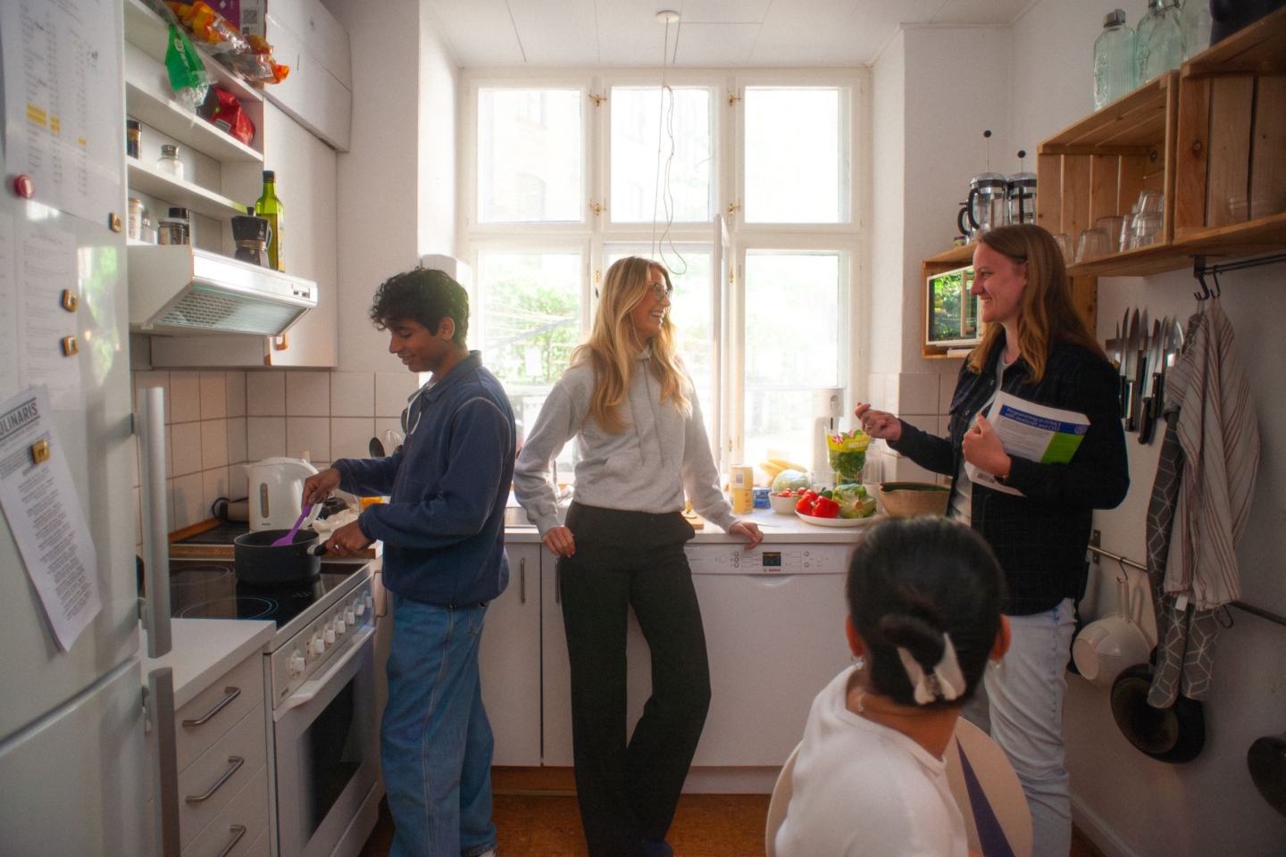 vidvinkel billede af et køkken med fire personer. Tre står i baggrunden foran et stort vindue. En sidder i forgrunden med ryggen vendt til.